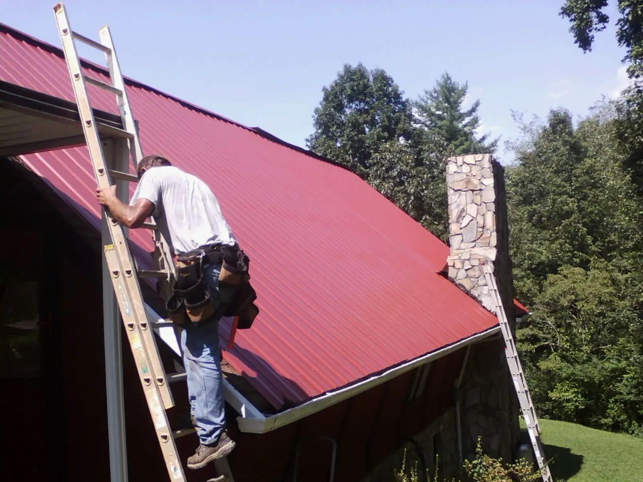 Roofer on a ladder beside a red metal roof and stone chimney — Soffit & Fascia Repair by Roofing Brothers in Clairfield